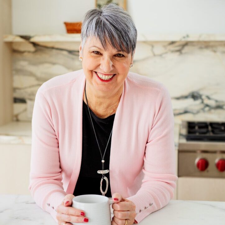 Image of Cathy Turalinski, grey hair, with pink cardigan and black blouse underneath. She is looking at camera smiling, leaning over a countertop holding a mug of coffee.